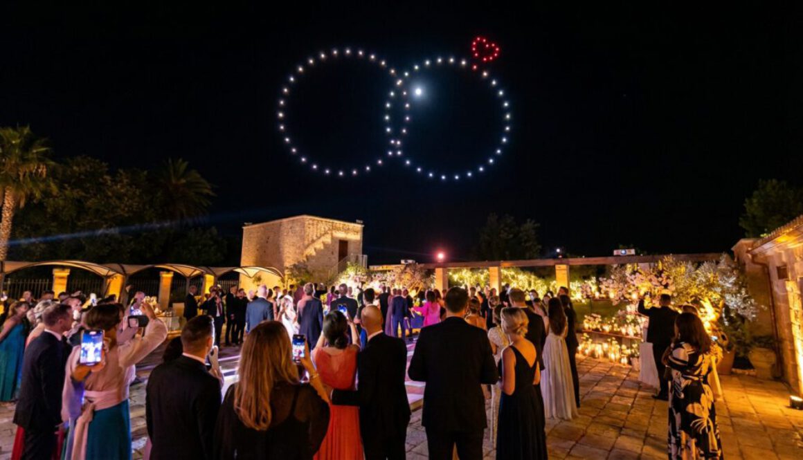 Gäste einer Abendhochzeit blicken in den Nachthimmel, wo eine Drohnenshow zwei riesige, leuchtende Eheringe mit einem kleinen roten Herz formt.