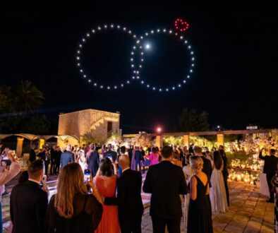 Gäste einer Abendhochzeit blicken in den Nachthimmel, wo eine Drohnenshow zwei riesige, leuchtende Eheringe mit einem kleinen roten Herz formt.